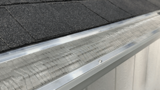 Close-up of a stainless steel micro-mesh gutter guard, made in USA, installed along the edge of a roof with dark shingles to keep debris out of the gutter below.